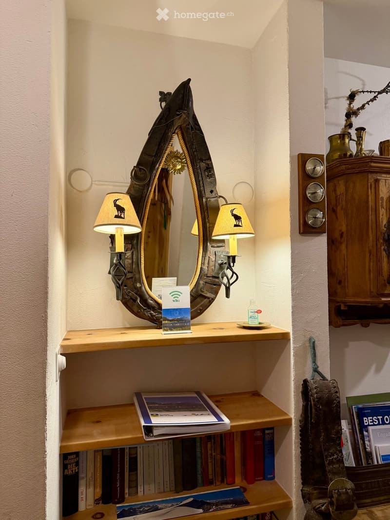 A cozy corner with a wooden shelf holding books, a mirror with decorative lamps, a weather station, and a small cabinet with decorative items.