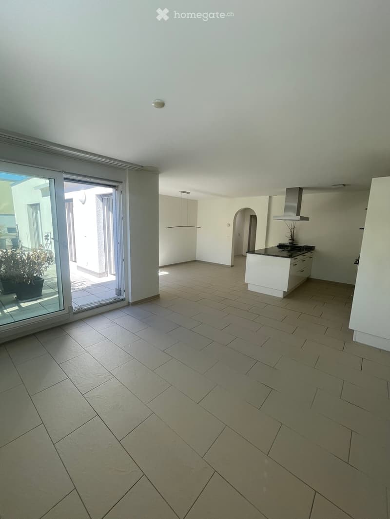 Empty room with tiled floor, white walls, and a sliding glass door leading to an outdoor area. A small kitchen area is visible in the background with cabinets and a countertop.