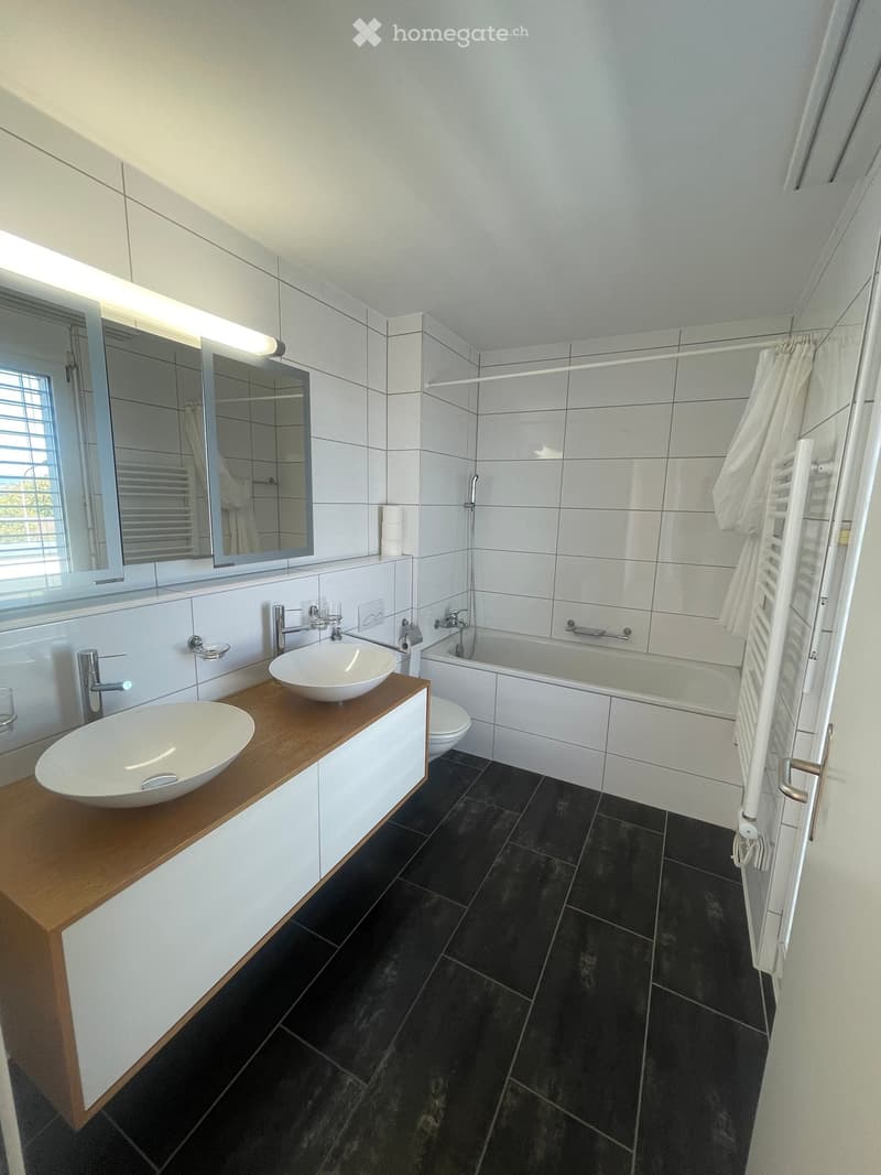 Modern bathroom with white tiles, dark floor tiles, double sink vanity, bathtub, large mirror, and towel rack.