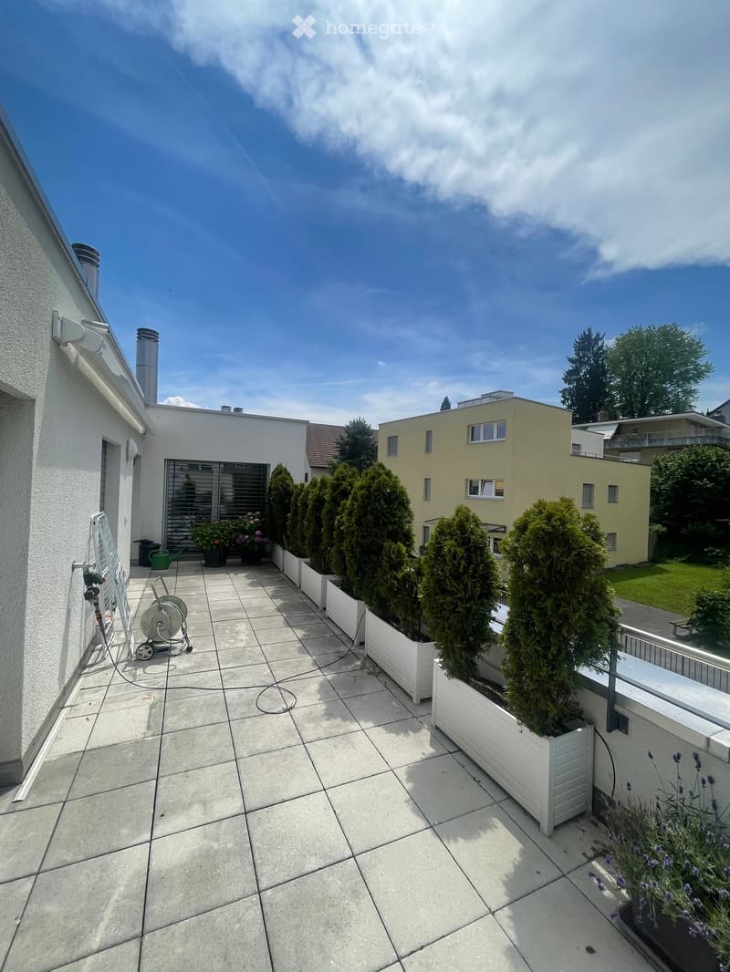 A tiled patio area with white planters containing tall, conical evergreen trees, a small garden hose reel, and a green watering can. The patio is bordered by a white railing and has a view of neighboring houses and greenery.