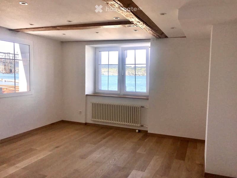 Empty room with wooden flooring, white walls, exposed wooden beams on the ceiling, and two windows offering a view of the sea.