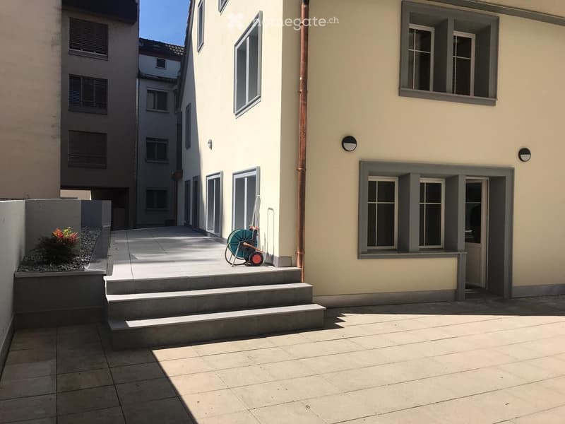 Courtyard area with tiled flooring, steps leading up to a building entrance, small garden bed with plants, and a hose reel.