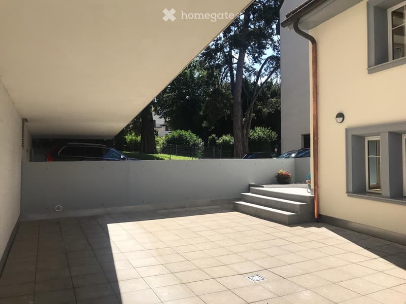 Outdoor patio area with tiled flooring, steps leading up, covered roof section, and parked cars visible in the background.