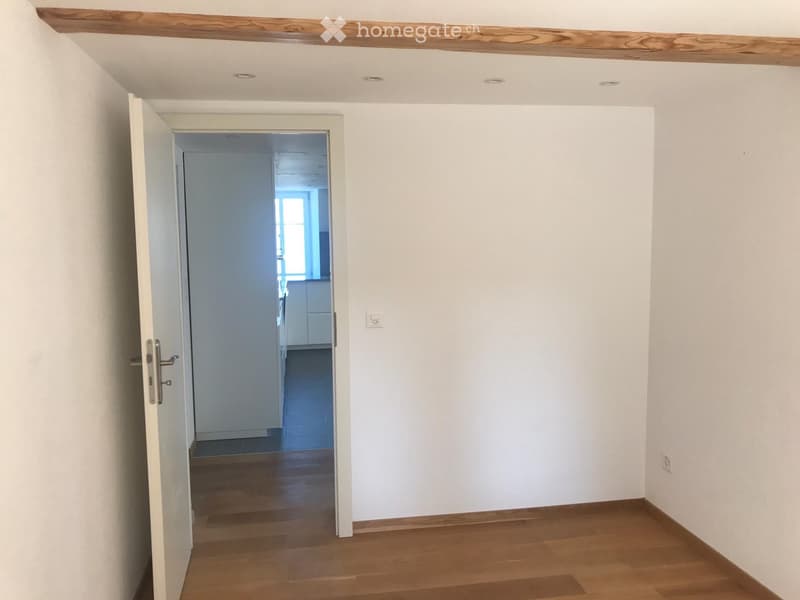 Empty room with wooden flooring, white walls, and an open door leading to another room with white cabinets and a window.