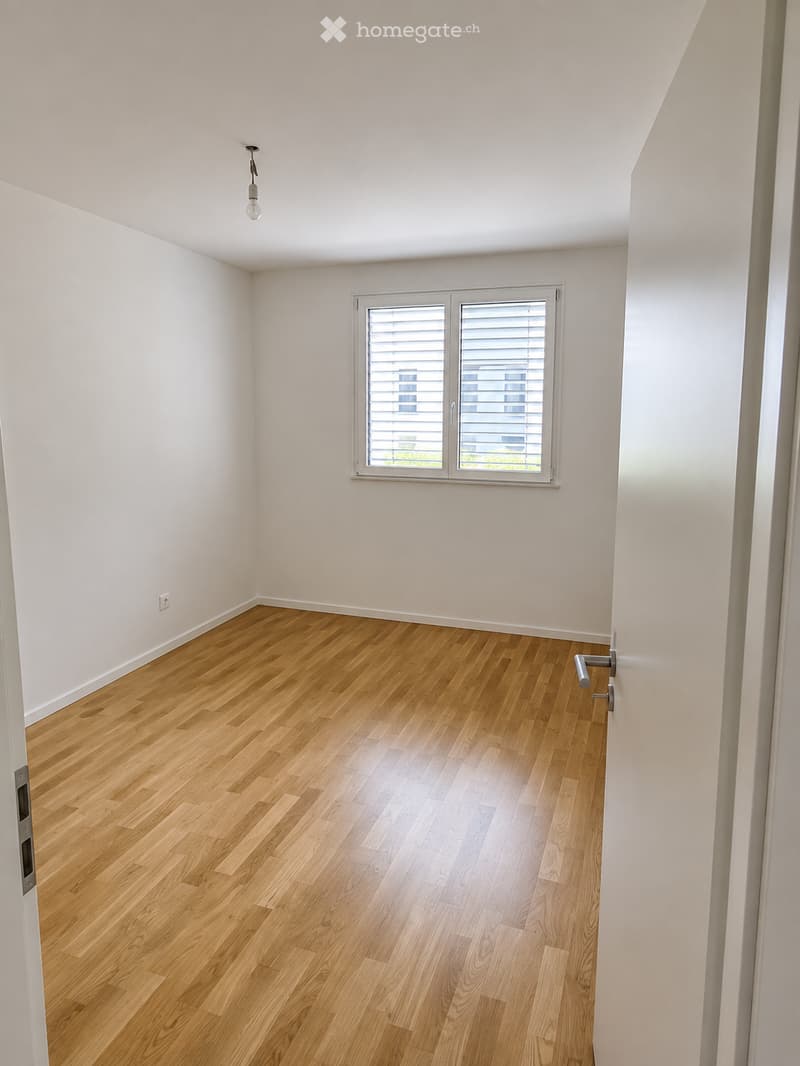 Empty room with light wooden flooring, white walls, and a single window with blinds. The room has a simple, modern design with a visible light fixture on the ceiling and an electrical outlet on the wall.