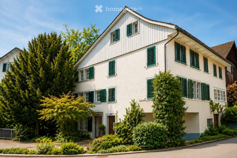 Two-story white house with green shutters, surrounded by trees and shrubs, with a driveway and entrance.
