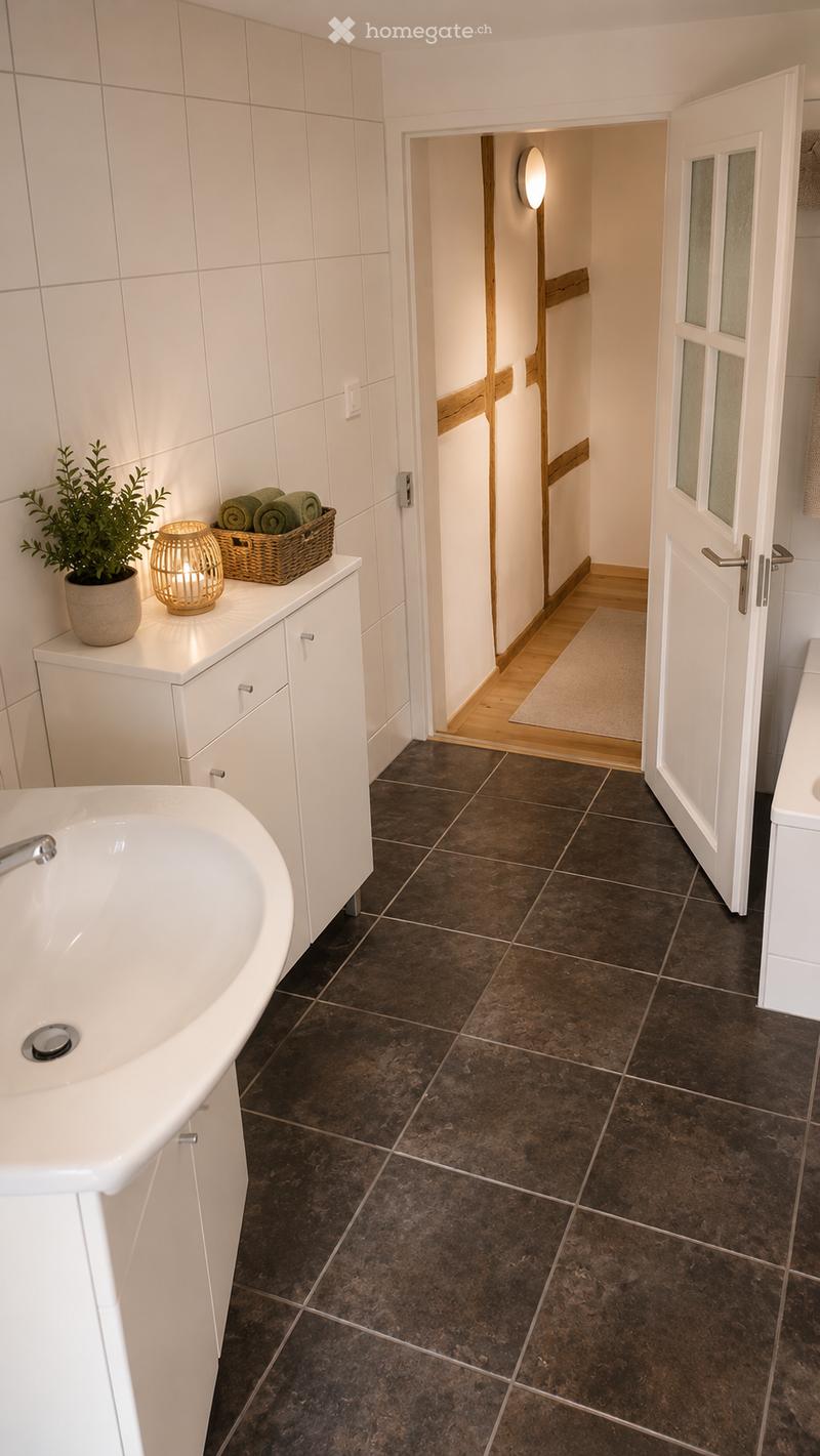 Modern bathroom with dark brown tiles, white cabinetry, sink, wicker basket with towels, small plant, and a wicker lamp on the counter. An open doorway leads to a hallway with wooden flooring.