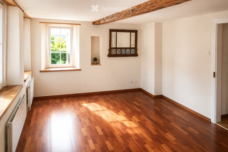 Empty room with wooden flooring, white walls, and large windows allowing natural light. Features include a built-in shelf with a small plant and a decorative mirror above it.