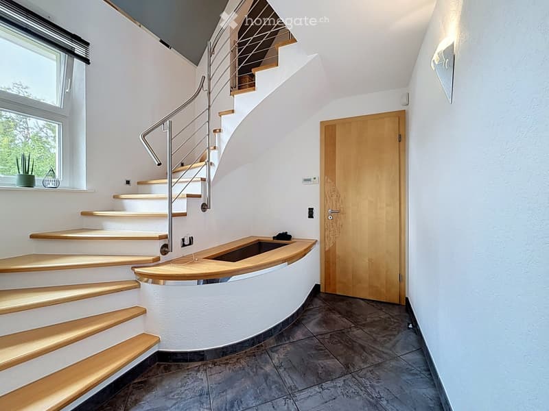 Spiral staircase with wooden steps and metal railing, white walls, large window, wooden door, and small shelf with decorative items.