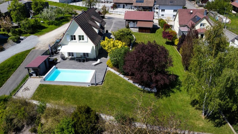 Aerial view of a modern house with a swimming pool, surrounded by a well-maintained lawn and trees. The house features a white exterior with a dark roof and a covered patio area with outdoor furniture.