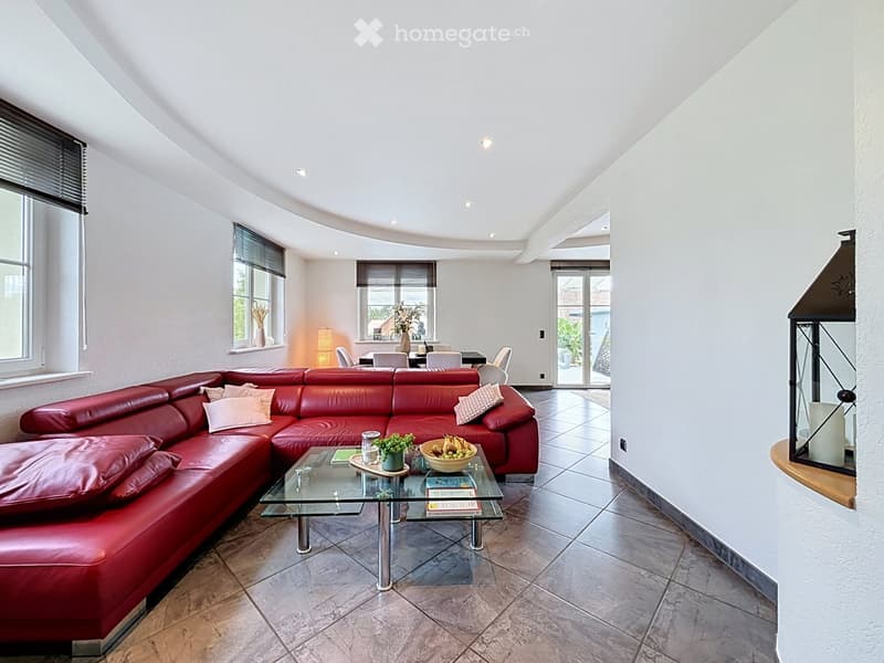 Modern living room with a red leather sectional sofa, glass coffee table, dining area with chairs and table, large windows with blinds, and decorative vases on sills.