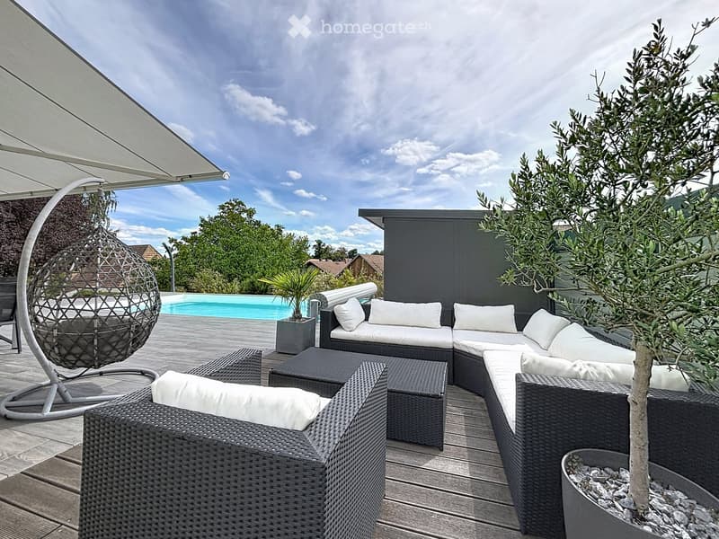 A modern outdoor patio with a wicker sofa, chairs, a hanging egg chair, a small table, and a potted plant, overlooking a swimming pool and garden area.