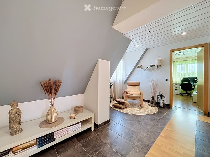 A modern, well-lit living space with a slanted ceiling and skylight. Features include a white table with books and decorative items, a beige armchair, a round rug, and a small sitting area with a lamp and decorative items.