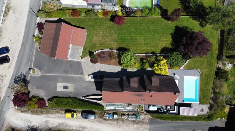 Aerial view of a two-story house with a red-tiled roof, surrounded by a well-maintained lawn, trees, and a swimming pool. There is a driveway with several parked cars and a garage visible.