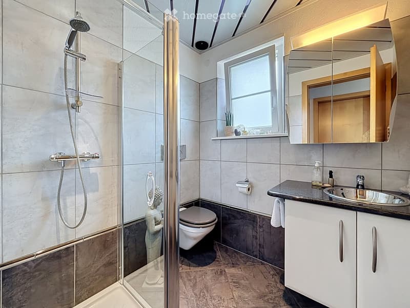 Modern bathroom with a glass-enclosed shower, toilet, sink, and mirrored cabinet. Features a sleek design with grey tiles and a small window above the sink.