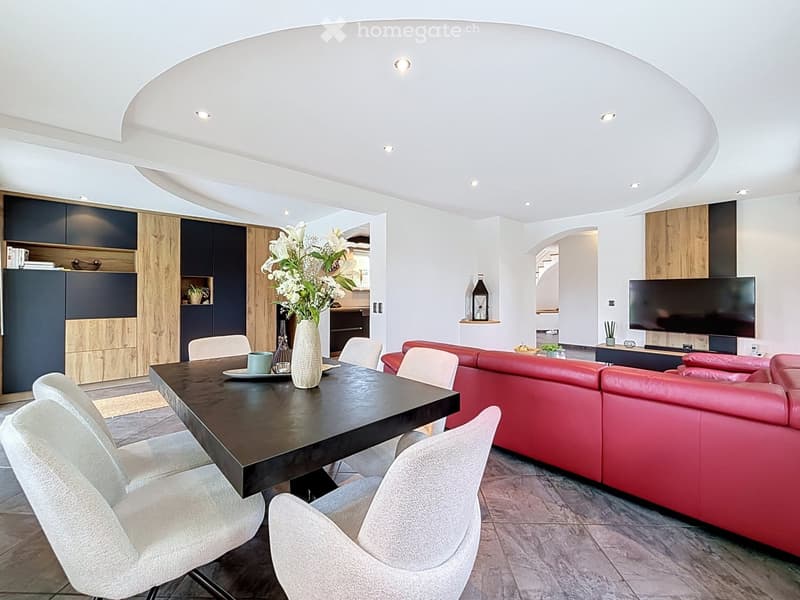 Modern living room with a red leather sofa, a dark wooden dining table, white chairs, a vase with white flowers, and a flat-screen TV on a stand.