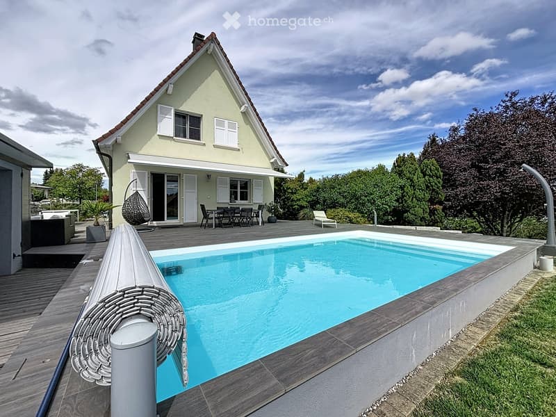 Outdoor area with a swimming pool, wooden deck, outdoor furniture, and a modern house with white shutters and a sloped roof.
