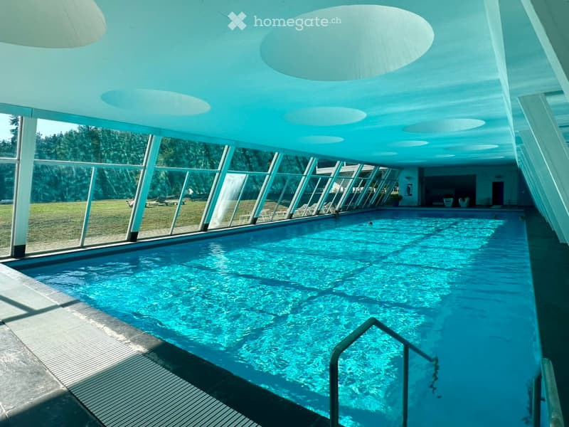 Indoor swimming pool area with large glass walls, surrounded by outdoor seating and greenery.
