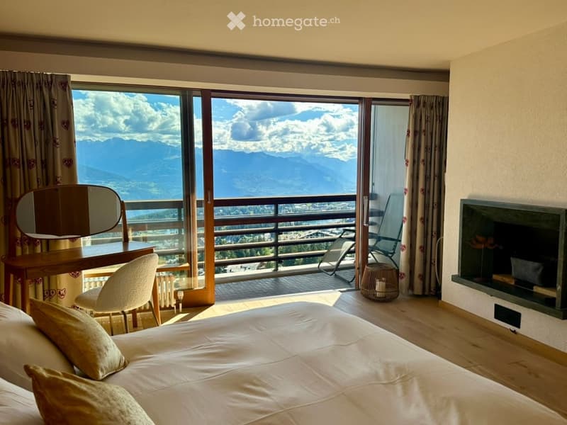 A modern bedroom with a large bed, pillows, a chair, a mirror, a table, and a fireplace, offering a scenic mountain view through large windows and a balcony.