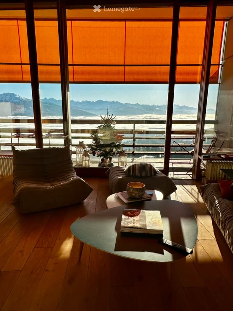 Bright living room with large windows offering a scenic mountain view, featuring a sofa, coffee table, armchair, and a small table with books and a remote control.