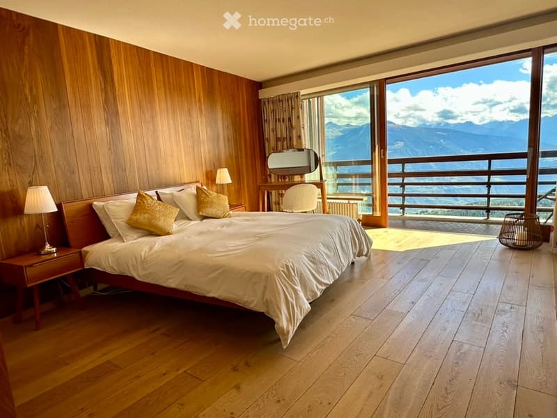 Spacious bedroom with wooden walls and floors, large bed with white linens and gold pillows, nightstand with lamp, vanity with mirror, and a view of mountains through large windows.