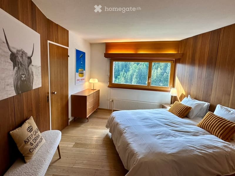 Cozy bedroom with wooden walls and flooring, a large bed with pillows and blankets, a bench with a cushion, a nightstand with a lamp, a radiator, a wooden cabinet, and artwork on the walls.