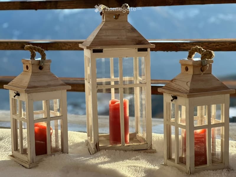 Three wooden lanterns with red candles placed on a snowy surface, likely a balcony or terrace, with a wooden railing and a blue sky in the background.