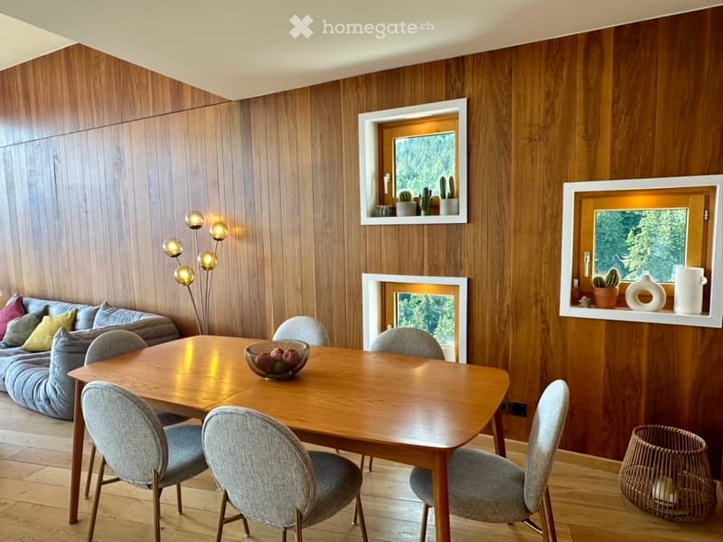 A modern living and dining area with wooden walls, a grey sofa with colorful cushions, a wooden dining table with six chairs, a floor lamp, and decorative items on the windowsills.