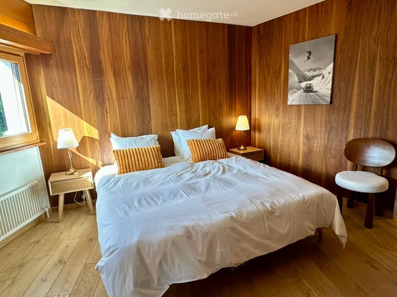 Cozy bedroom with wooden walls and floor, a large bed with white bedding and orange pillows, two nightstands with lamps, a wooden chair, and a stool.
