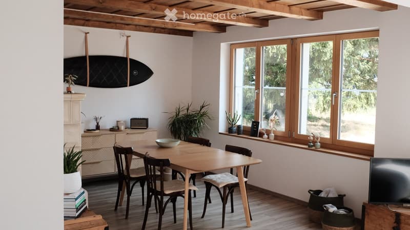 Bright dining area with wooden table and chairs, cabinet with decorative items, surfboard on wall, and large windows with plants on the sill.