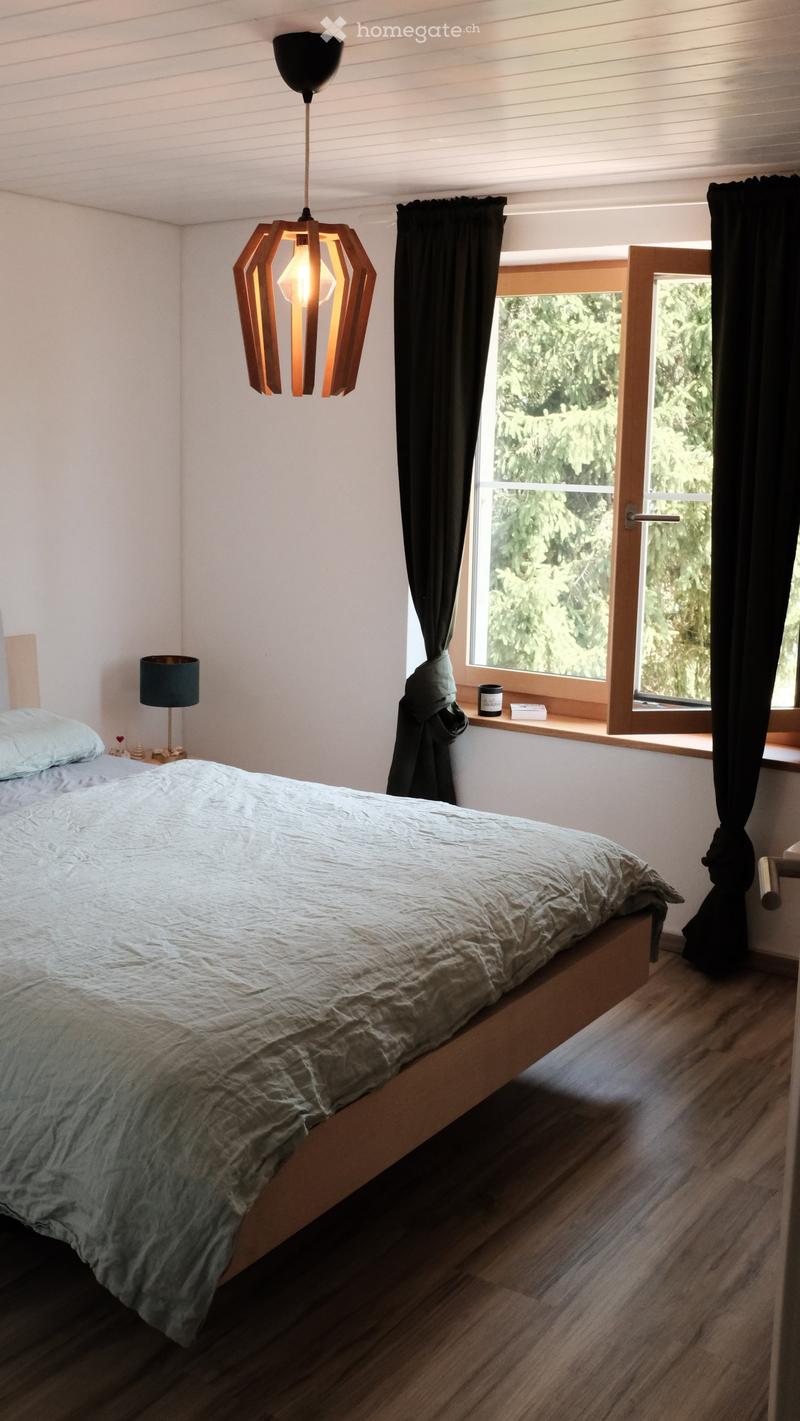Cozy bedroom with a wooden bed, light-colored bedding, a pendant light fixture, a table lamp, and a window with dark curtains overlooking a green landscape.