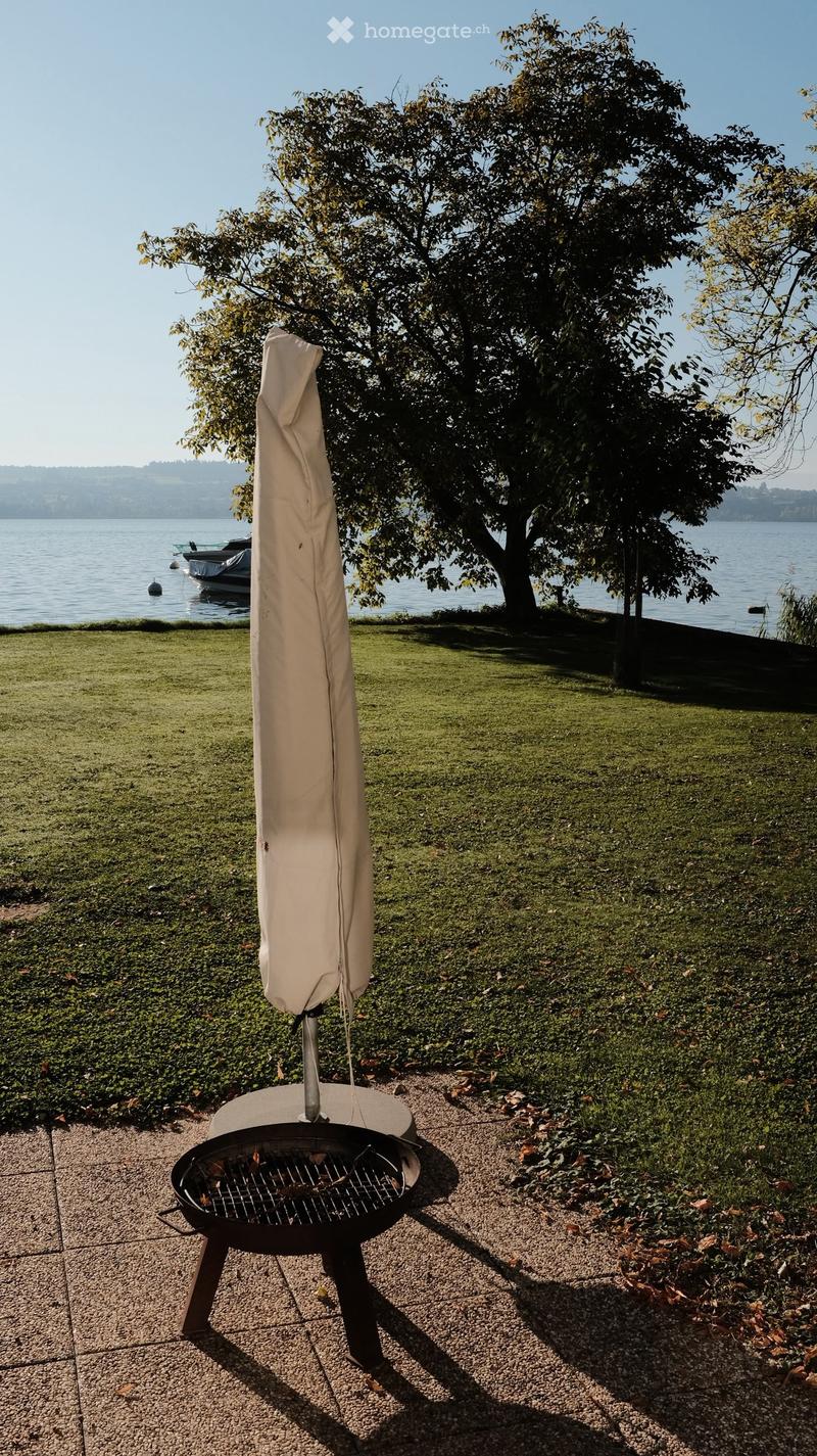 Outdoor garden area with a grill, umbrella, and a view of a lake with a boat in the background.