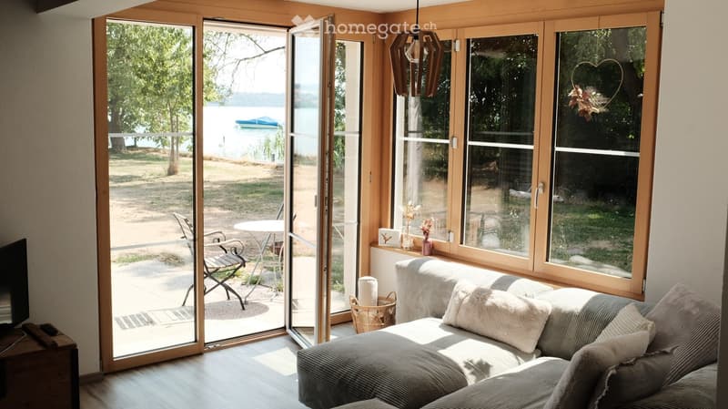 Bright living room with large sliding glass doors opening to a patio, overlooking a lake. Features a grey sofa with white pillows, a small round table, a wicker basket, and a television on a wooden stand.