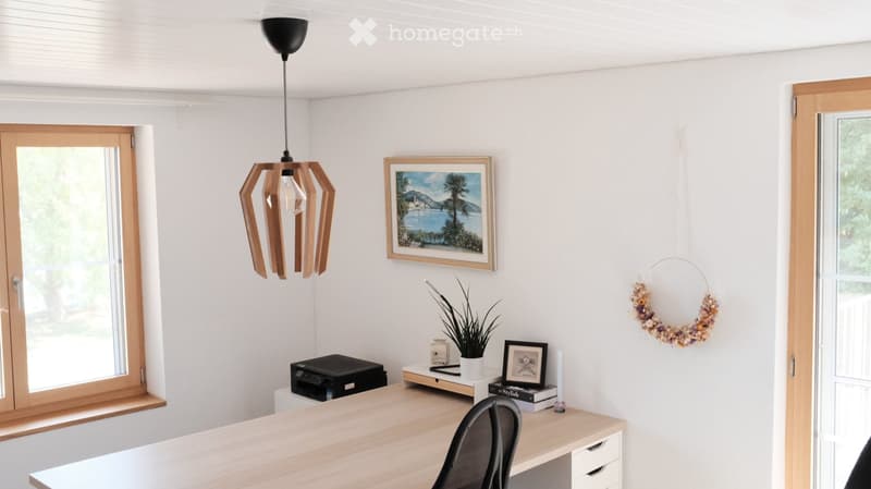 Bright, modern room with white walls, wooden desk, black chair, hanging pendant light, and decorative items including a framed picture, a wreath, and a small plant.