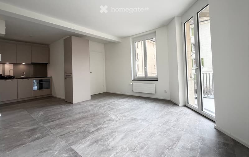 Open-plan space with modern kitchen area featuring cabinets, sink, and oven, connected to a living area with large windows and a sliding glass door leading to a balcony.