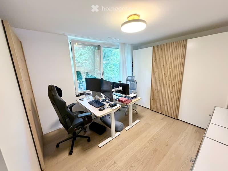 A modern, well-lit office space with a large window, a standing desk, ergonomic chair, multiple computer screens, and various office supplies.
