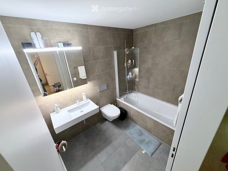 Modern bathroom with a white sink, toilet, bathtub, and large mirror. Grey tiled walls and floor, with a small shelf holding toiletries.