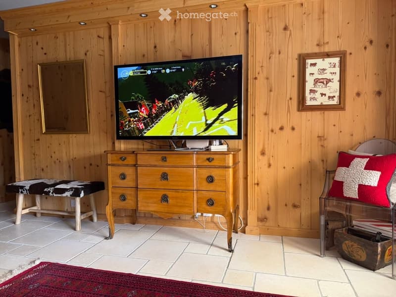 A cozy living room with wooden walls, a television on a chest of drawers, a stool, a chair with a red cushion, and a framed picture on the wall.