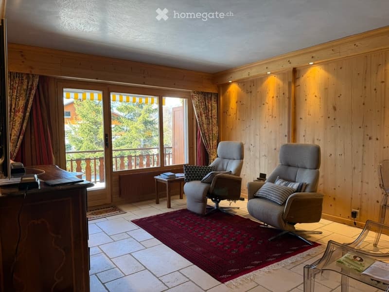 A cozy living room with wooden walls, large windows leading to a balcony, two armchairs, a small table, and a laptop on a wooden cabinet.