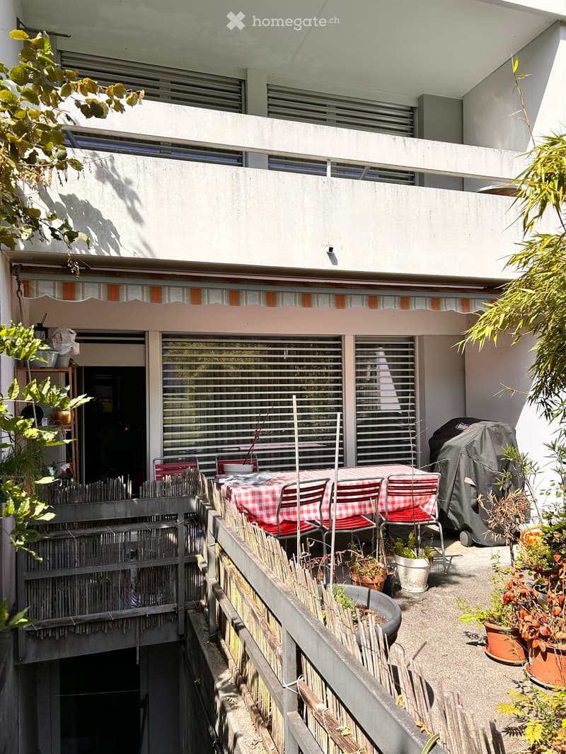Balcony with a red and white checkered tablecloth, chairs, potted plants, and a covered grill. The balcony has a metal railing and is partially shaded by a striped awning.
