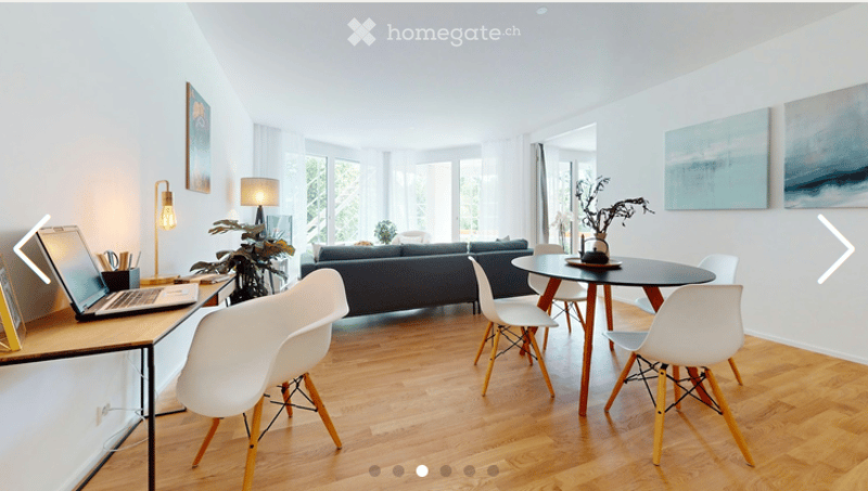 Modern living room with a dark blue sofa, white chairs, a round dining table, a laptop on a desk, and decorative plants and artwork on the walls.
