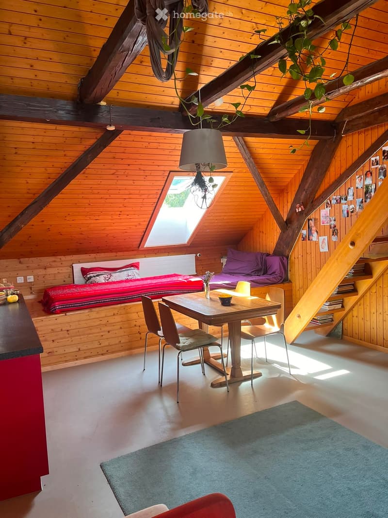 Cozy attic room with wooden walls and exposed beams, featuring a sofa, dining table, chairs, and a lamp.