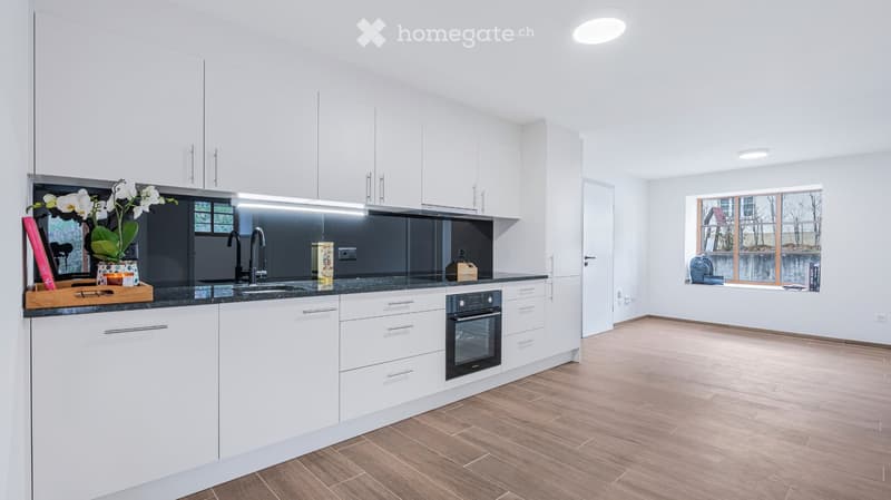 Modern kitchen with white cabinets, dark countertops, stainless steel sink and faucet, built-in oven, and a small seating area with a window view.
