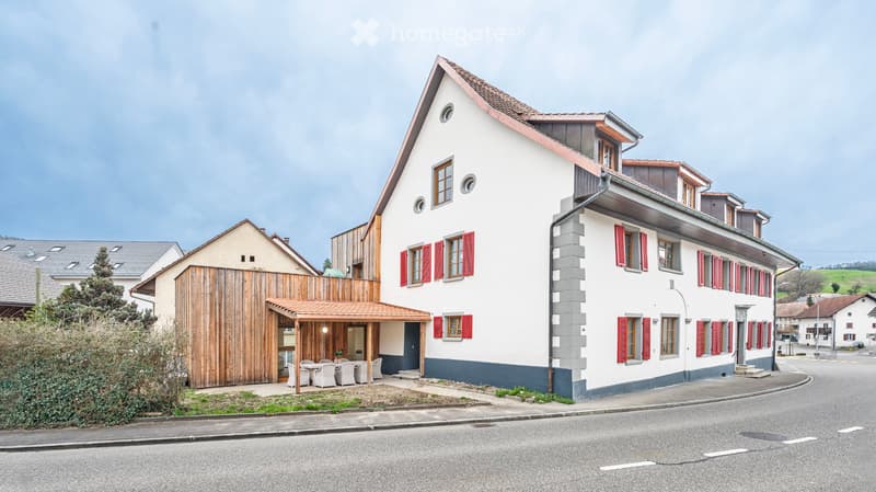 Two-story house with white walls, red shutters, and a wooden balcony area with outdoor seating.