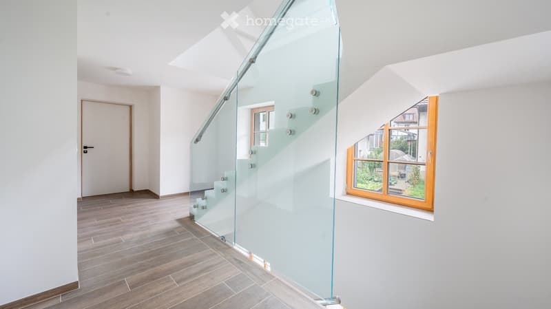 Modern hallway with wooden flooring, white walls, and a glass staircase leading upwards. Features large windows with wooden frames and a door on the left side.