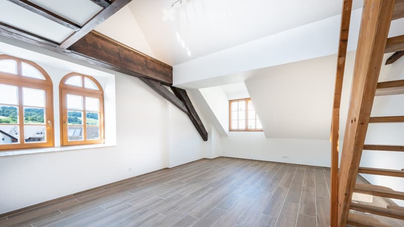 Empty attic room with wooden beams, white walls, and large windows offering a view of the landscape. The floor is covered with light-colored wooden tiles.