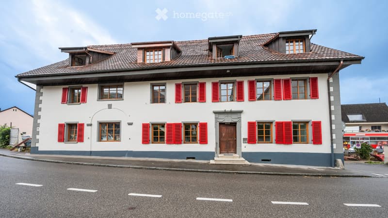 Two-story house with red shutters, white walls, and a tiled roof. Features a central wooden door and multiple windows.