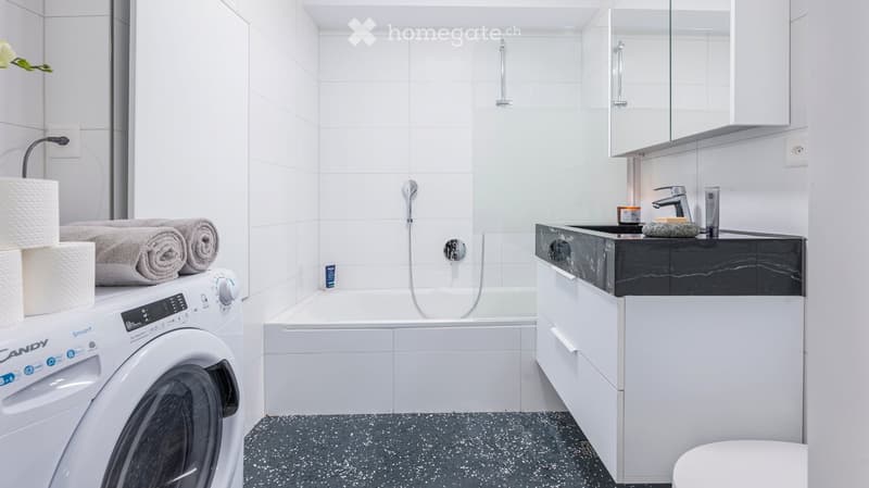 Modern bathroom with a washing machine, bathtub, sink, mirror, and neatly rolled towels on top of the washing machine.