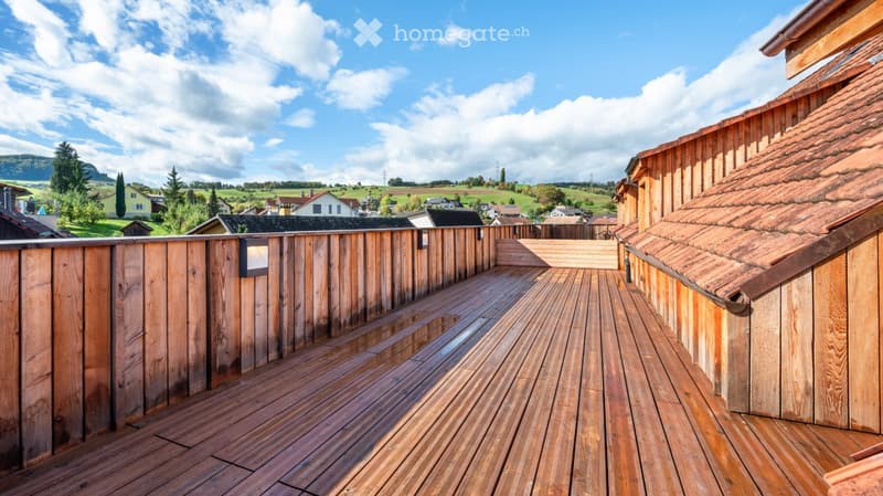 Wooden balcony with wooden decking, wooden railing, and a view of a rural landscape with houses and trees.