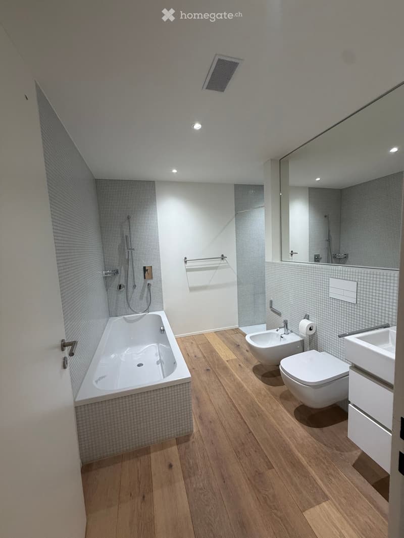 Modern bathroom with wooden flooring, a bathtub, toilet, sink, bidet, and a large mirror. The walls are tiled with a light grey pattern, and the space includes a shower area with a glass enclosure.